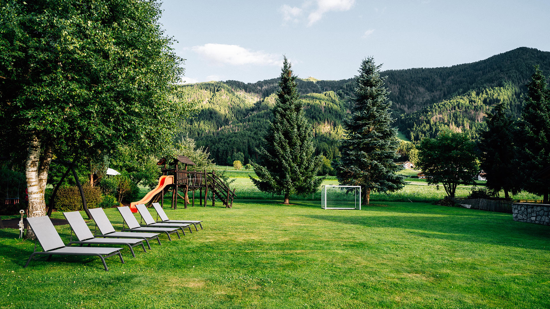 Ampio giardino dell'hotel con sedie a sdraio, parco giochi per bambini e porta da calcio di fronte a montagne verdi al Kronplatz.