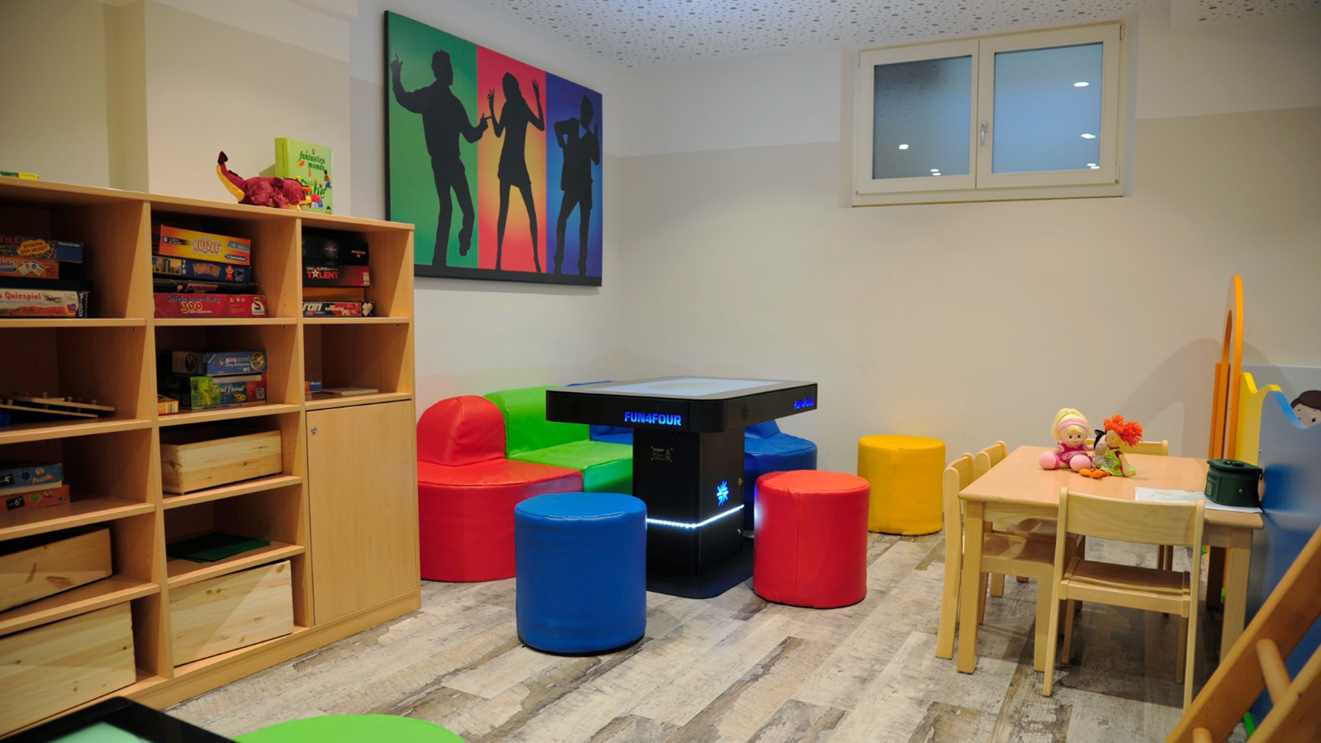 Luminosa area giochi all'Hotel Tannenhof con tavolo interattivo, sedute colorate, scaffale in legno pieno di giochi da tavolo e tavolo per bambini con bambole.