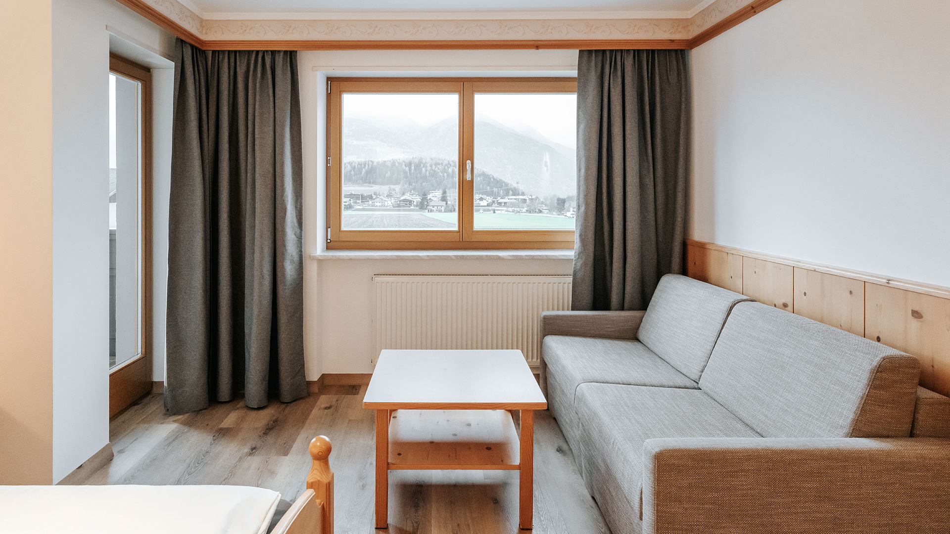 Accogliente camera matrimoniale nell'Hotel Tannenhof con pavimento in legno chiaro, divano, tavolo e vista sulle montagne del Kronplatz.
