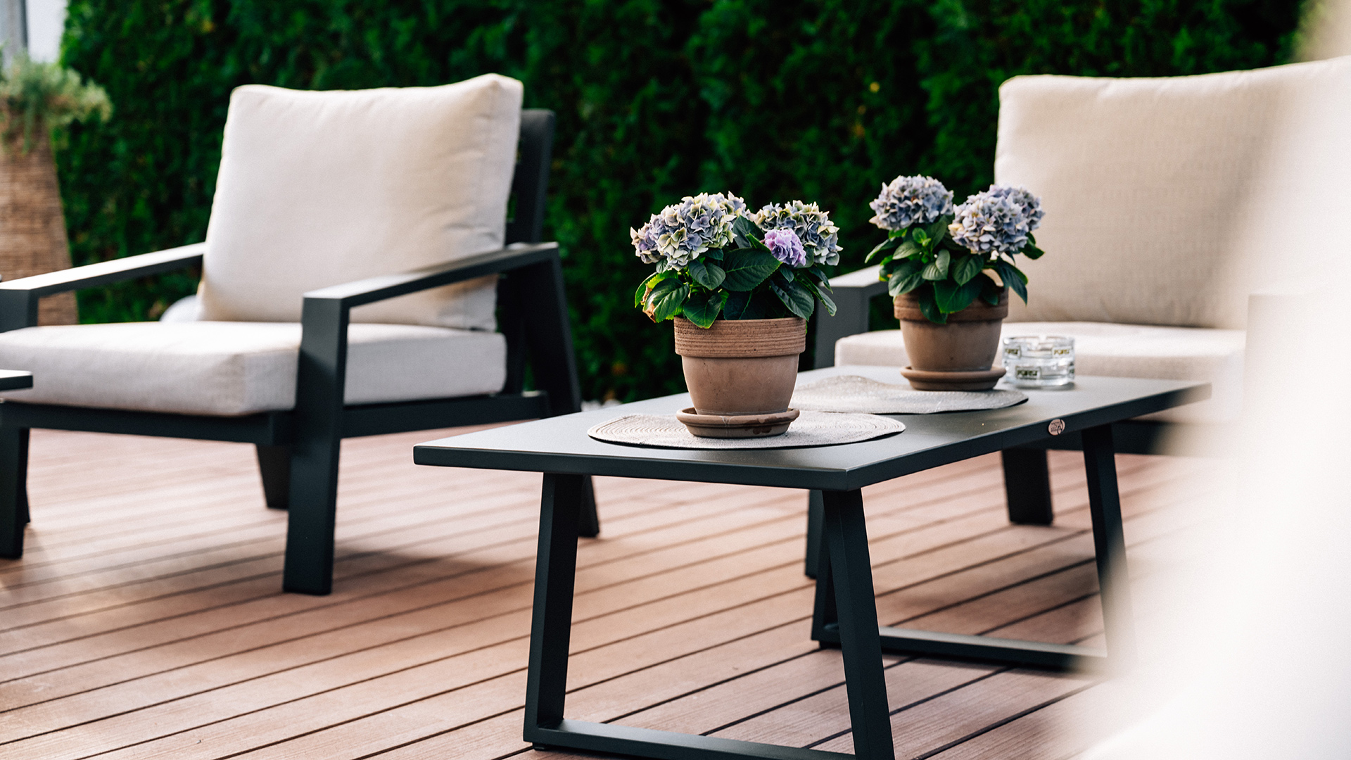 Due comode poltrone con tappezzeria chiara e un tavolo scuro con ortensie sulla terrazza in legno dell'Hotel Tannenhof.