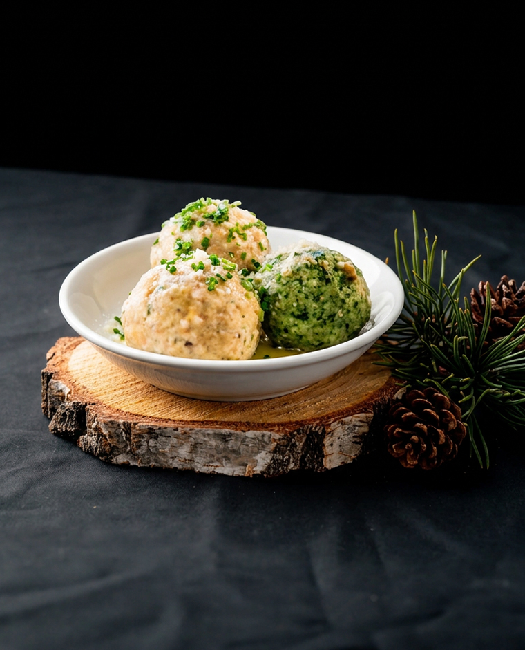 Tre Knödel Tris del Südtirol, due chiari e uno verde, in una ciotola bianca su un tagliere di legno.
