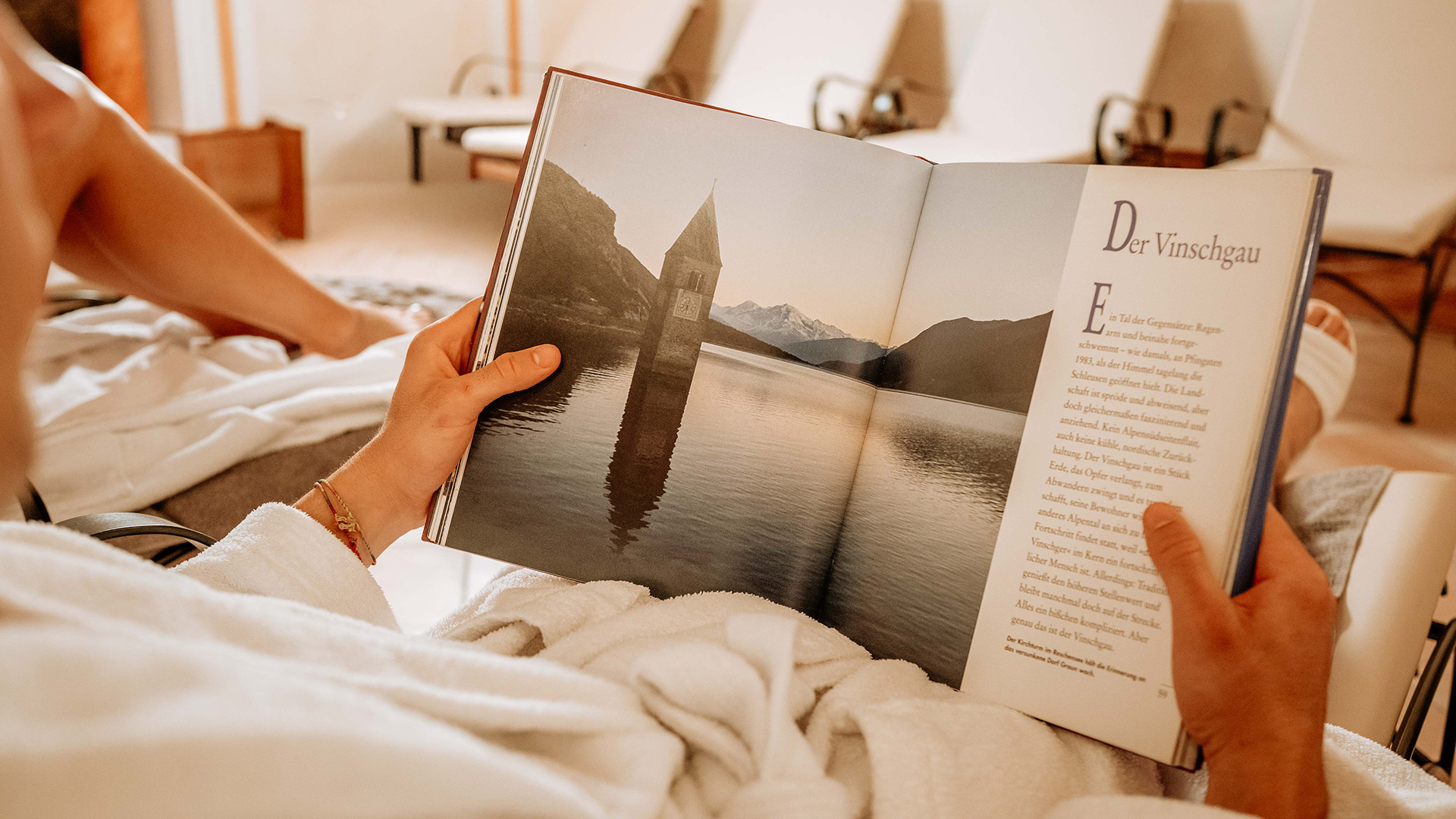 Eine Person entspannt im Bademantel auf einer Liege und liest ein Buch.