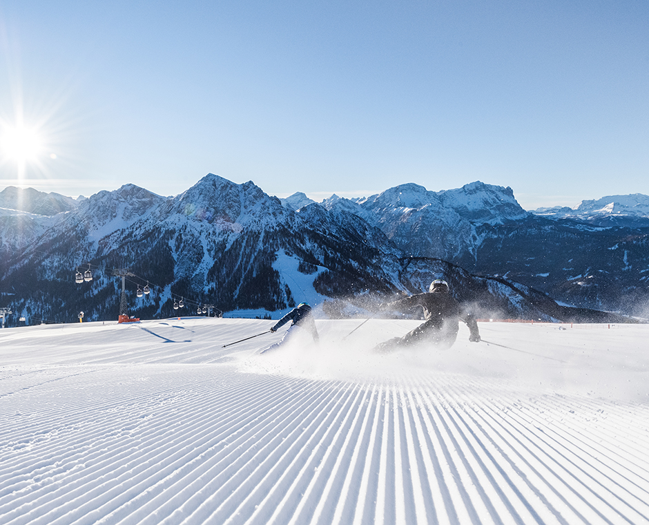Zwei Skifahrer carven dynamisch auf einer frisch präparierten Piste am Kronplatz mit verschneiten Bergen im Hintergrund.