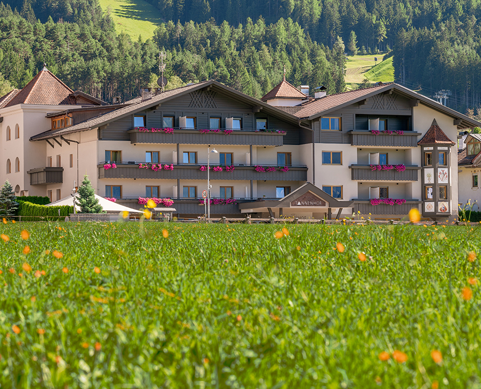 Das Hotel Tannenhof am Kronplatz in Südtirol, umgeben von grünen Wiesen mit Blumen und bewaldeten Bergen.