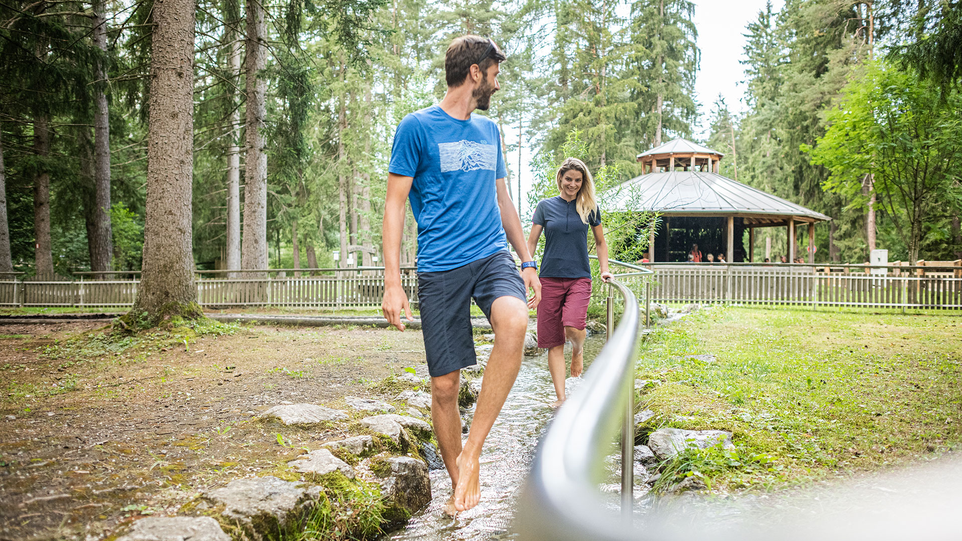 Ein Paar genießt eine Kneipp-Anwendung im Bach, umgeben von Wald und einem Pavillon, ideal für den Sommerurlaub am Kronplatz.