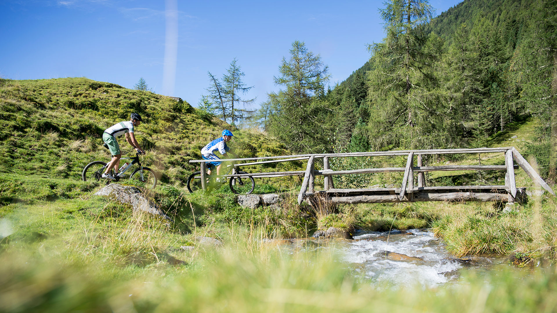 Zwei Mountainbiker auf einem sonnigen Trail am Kronplatz, einer überquert eine Holzbrücke über einen Bach.