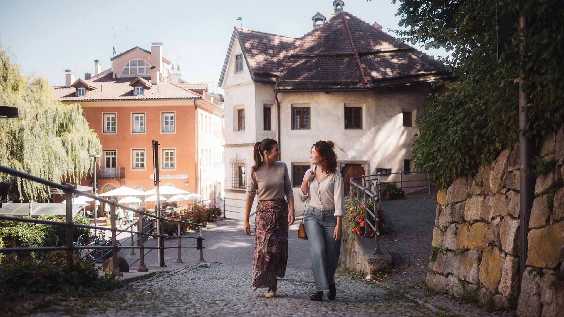 Zwei Frauen spazieren lächelnd durch eine charmante Gasse in einem Südtiroler Dorf, ideal für Sommerurlaub.