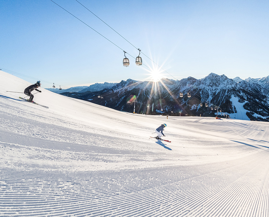 Two skiers on a perfectly groomed slope at Kronplatz, with gondolas and snow-covered mountains in the sunlight.