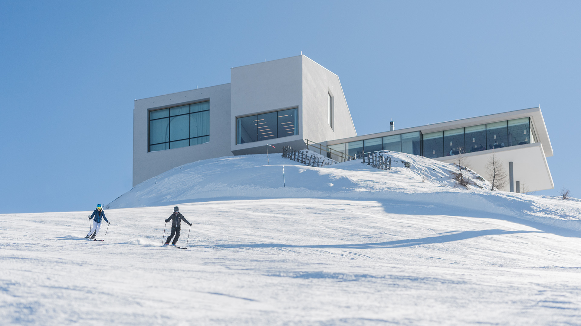 Zwei Skifahrer auf der Piste am Kronplatz mit dem modernen Lumen Museum im Hintergrund, ein Highlight für Winterurlauber.