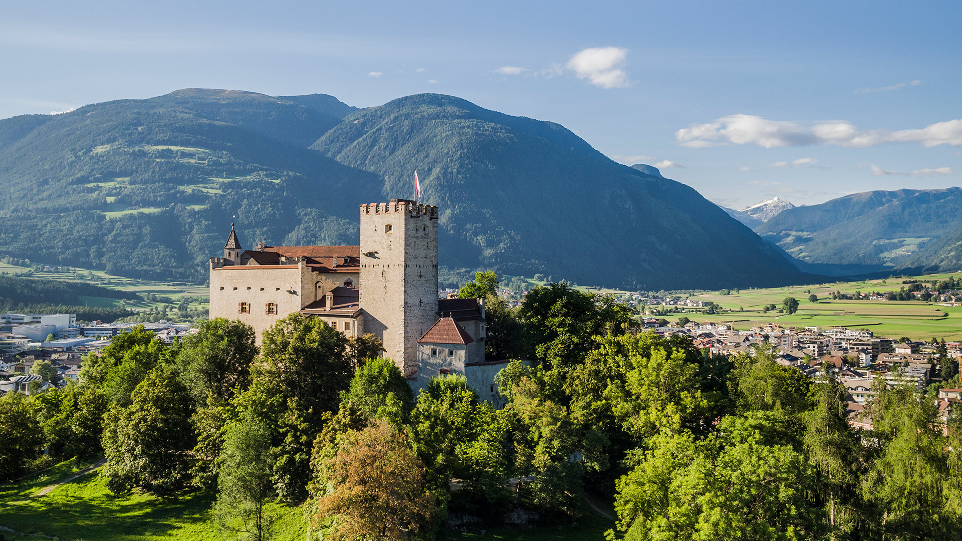 Historisches Schloss Bruneck auf einem grünen Hügel, umgeben von Bäumen, mit dem Pustertal und den Bergen im Hintergrund.