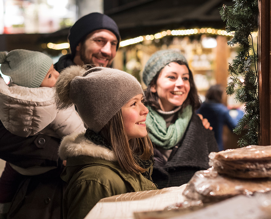 Eine lächelnde Familie mit zwei Kindern, warm gekleidet, schaut begeistert einen Weihnachtsmarktstand an.