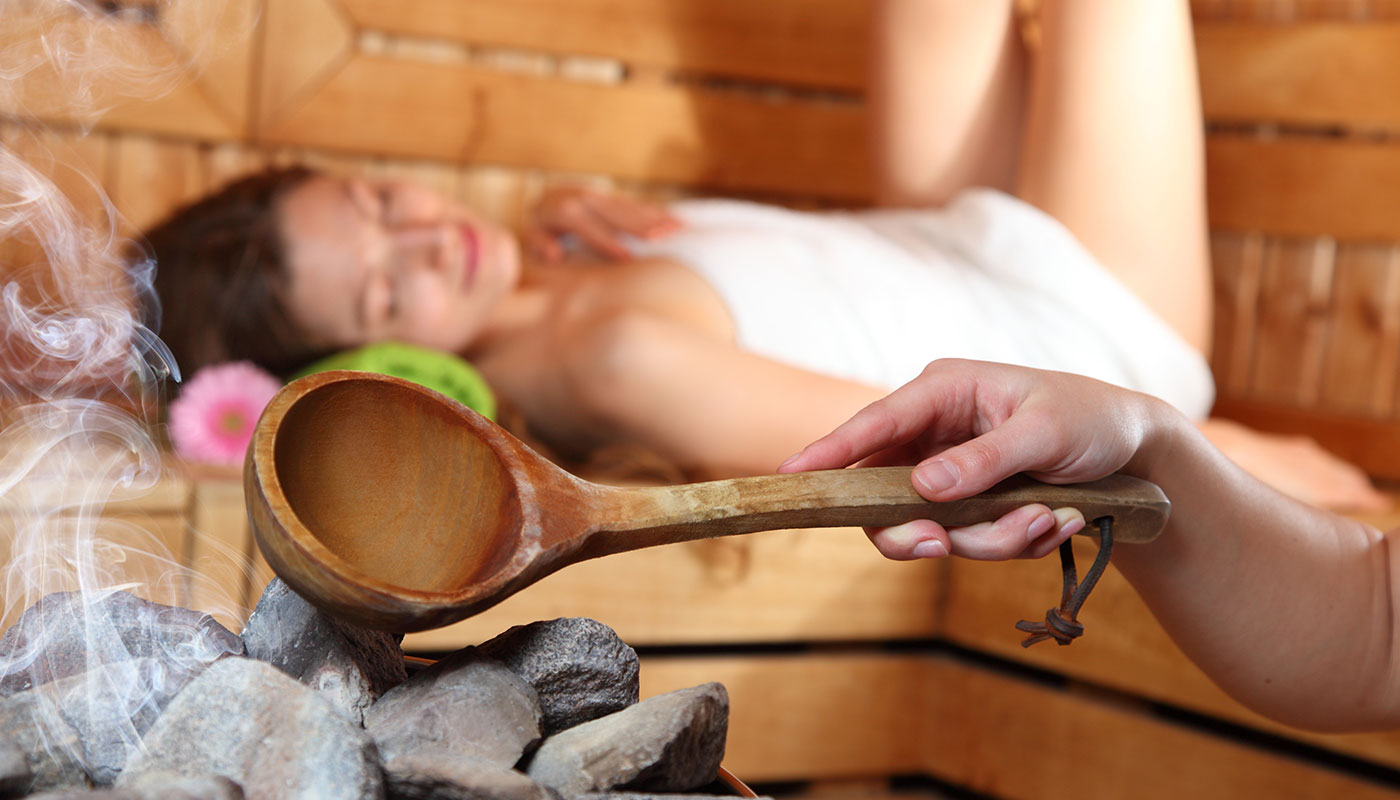 a young lady is lying relaxed in the sauna while an employee pours water over hot stones